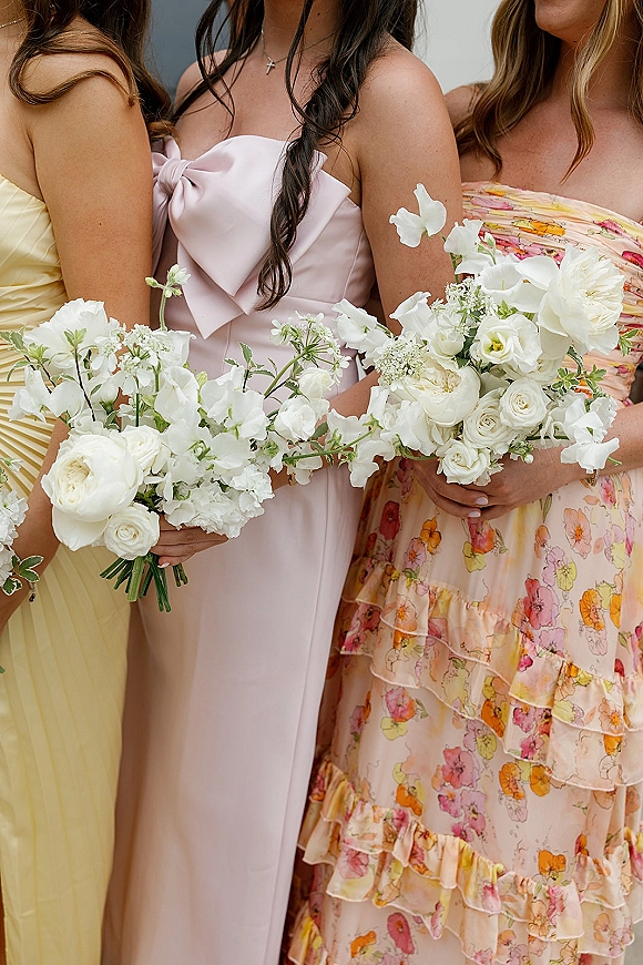 Bridesmaid bouquet of white bridesmaid bouquets with roses, sweet peas and greenery, held by bridesmaids in mismatched dresses by a neutral wall