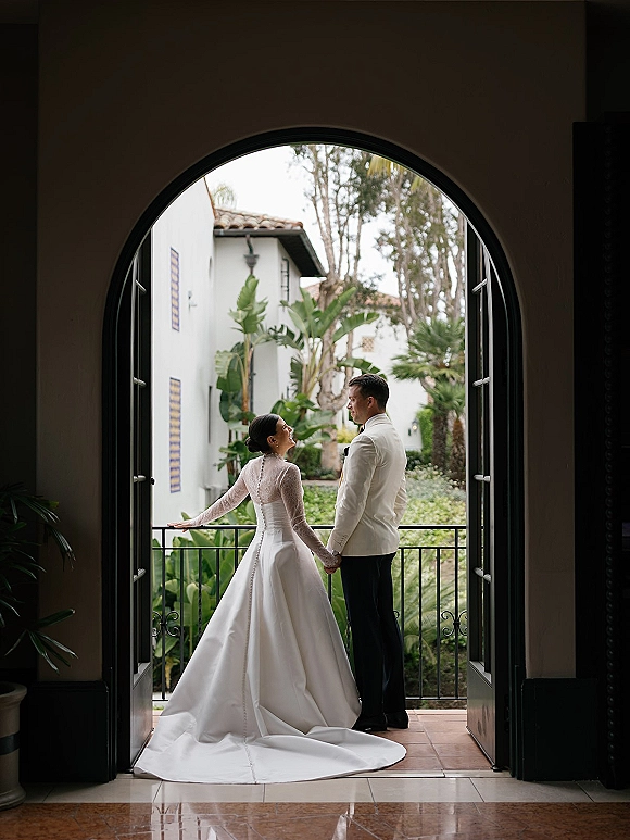 Couple portrait of bride and groom holding hands on a balcony, her lace long sleeve gown with train and veil in a Spanish-style courtyard garden