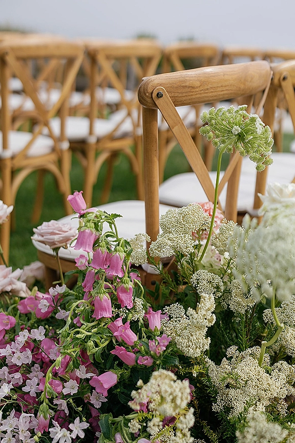 Ceremony aisle florals with grounded aisle flowers in pink bellflower and white wildflowers lining a grass lawn beside cross back chairs