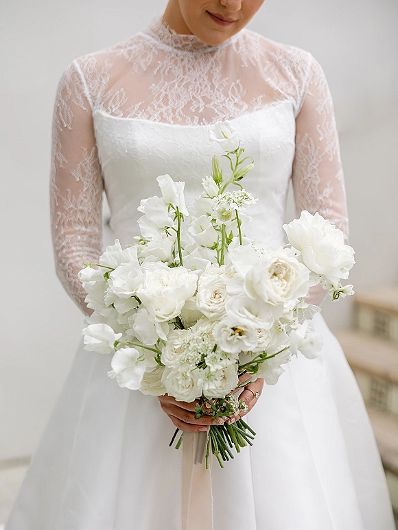 Bridal bouquet of white roses and sweet peas, tied with ribbon, held by a bride in a high-neck lace dress on stone steps