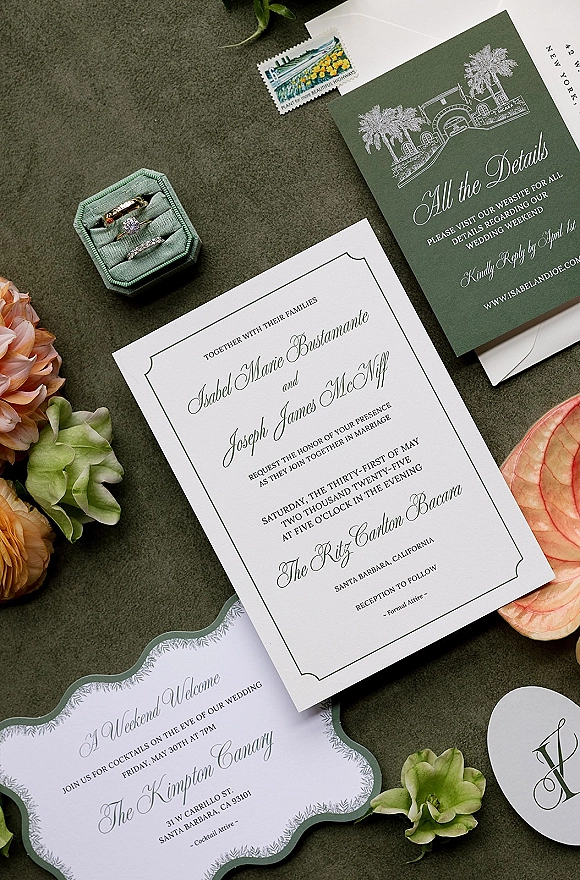 Invitation suite flat lay with wedding invitation cards, rings in a velvet box, flowers, and a postage stamp on green fabric surface