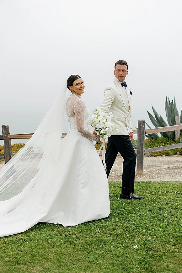 Couple portrait of bride and groom walking on a coastal lawn, bride looking back in long veil holding white bouquet with ocean horizon behind