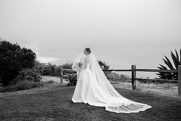 Bridal portrait in black and white of a bride in side profile with cathedral veil blowing in wind, holding bouquet by a wooden fence