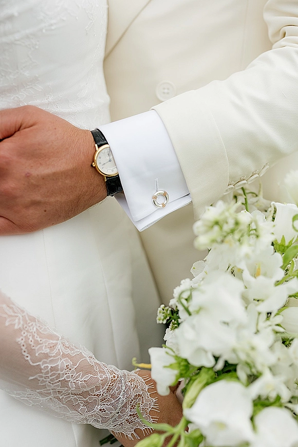 Wedding details of bride and groom hands, showing groom cufflinks, wristwatch, and bride lace sleeve holding a white bouquet on neutral backdrop