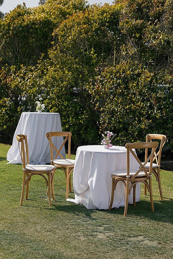 Outdoor cocktail tables with white tablecloths and pink bud vases, surrounded by wood cross back chairs on a sunny lawn by hedges