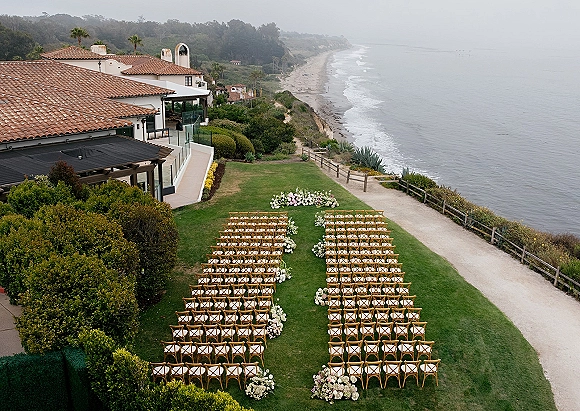 Ceremony setup with cross back chairs and aisle florals on a lawn overlooking the ocean shoreline and coastal cliffs under cloudy sky