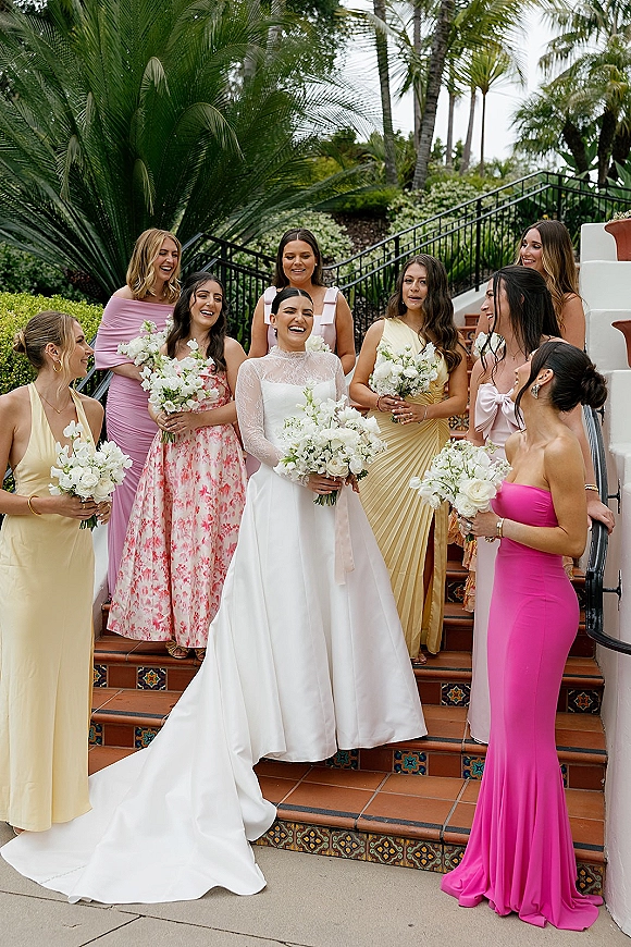 Bridesmaids group photo with bride and bridesmaids holding bouquets on a terracotta tile staircase, palm trees behind, gold hoop earrings