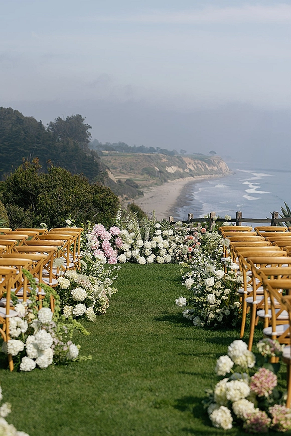Outdoor ceremony setup with hydrangea-lined aisle flowers and wood crossback chairs overlooking a cliffside ocean coastline and sky