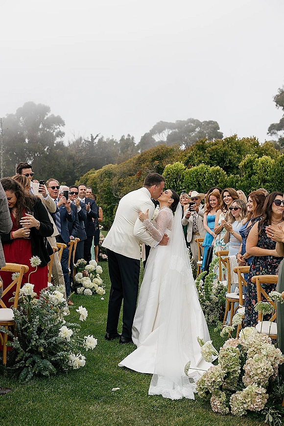 Ceremony kiss as bride in veil and lace dress and groom in suit walk between hydrangea aisle florals, guests with phones on garden lawn