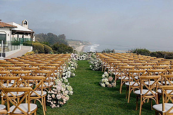 Outdoor ceremony setup with oceanfront wedding ceremony seating, wood crossback chairs and flower-lined aisle on a cliffside lawn by the coast