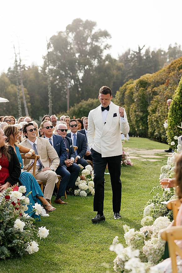 Groom walking aisle at an outdoor wedding processional in a white dinner jacket with black bow tie, passing hydrangea-lined chairs on a garden lawn