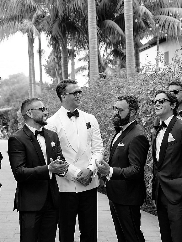 Groomsmen portrait of the groom and groomsmen in tuxedos with bow ties and sunglasses, lined up on a palm-lined walkway outdoors