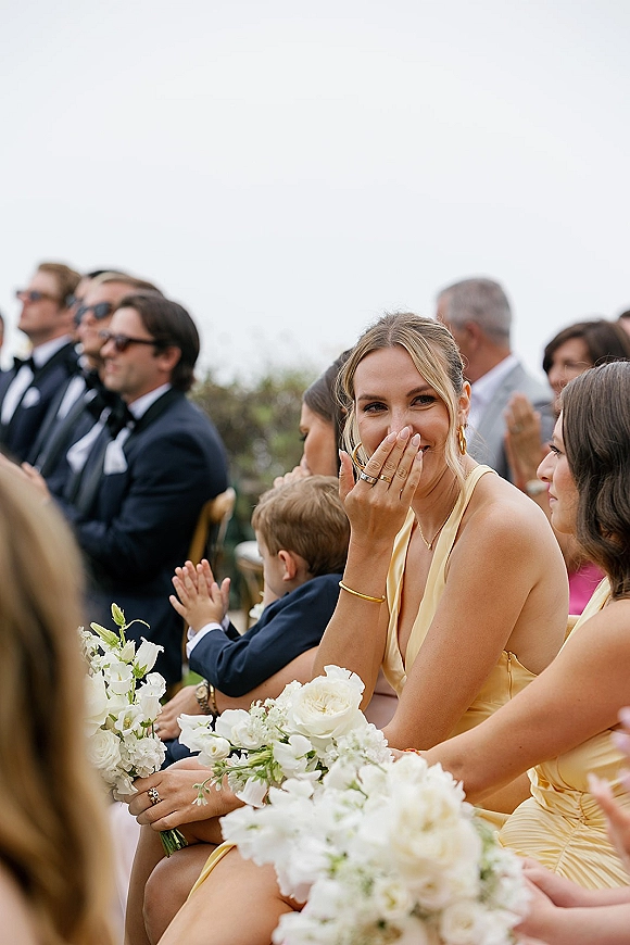 Wedding guest moment as bridesmaids in champagne dresses hold white bouquets, wearing gold hoops, seated outdoors as guests applaud