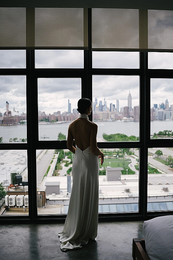 Bridal portrait of a bride by window in a backless wedding dress, facing floor-to-ceiling windows with city skyline and river view