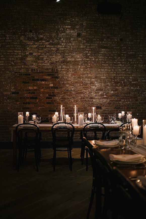 Reception tablescape with candlelit wedding reception taper and pillar candles in glass holders, set on wood tables before exposed brick wall