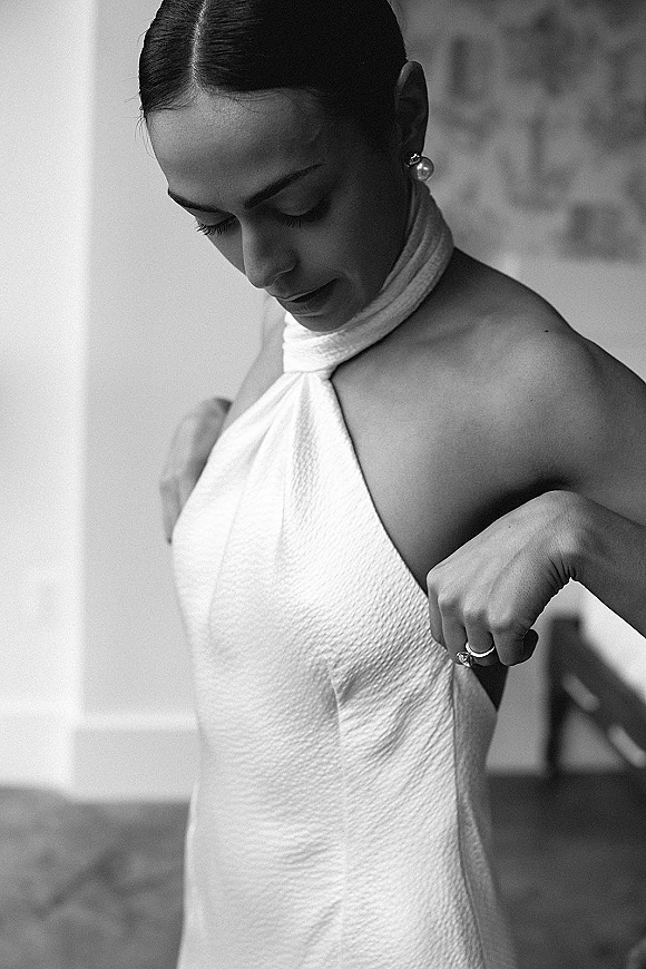 Bridal portrait of a bride getting ready, looking down while adjusting her halter wedding dress by window light in an indoor room