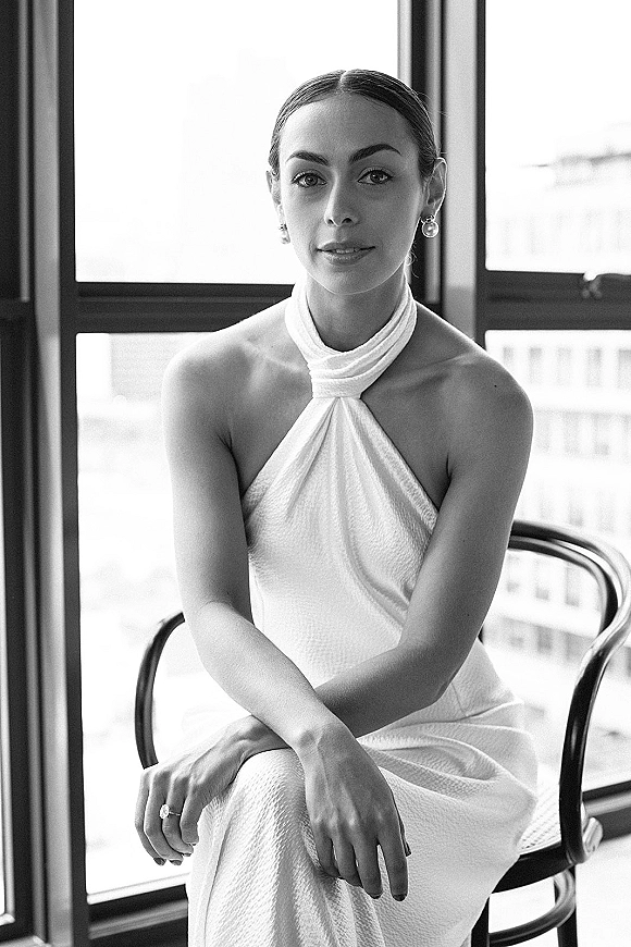 Bridal portrait of a bride in a halter wedding dress, seated by large windows with city buildings behind, pearl drop earrings visible
