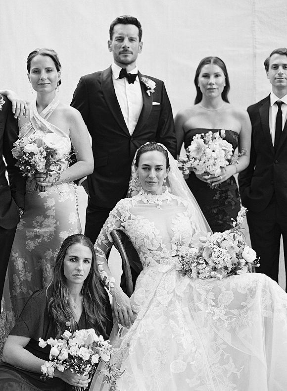 Wedding party portrait with bride seated in lace gown and veil, holding bouquet, as groom and attendants pose against a plain wall