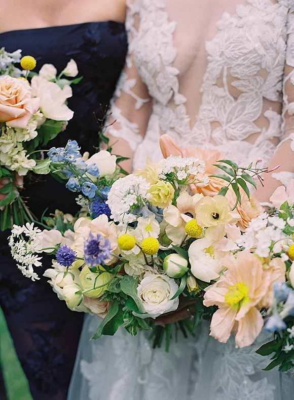 Bridal bouquet with wildflower wedding bouquet accents of blush roses, yellow billy balls, and blue blooms held against lace dress and tuxedo torso