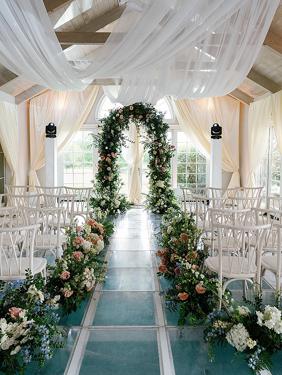 Ceremony setup for indoor wedding ceremony with a floral arch, glass aisle runner, and white chairs beneath arched windows