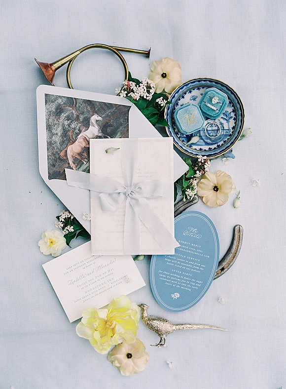 Invitation suite wedding invitation flat lay with blue and white cards, ribbon-wrapped envelope, rings on a vintage blue plate, and floral sprigs on a light blue surface