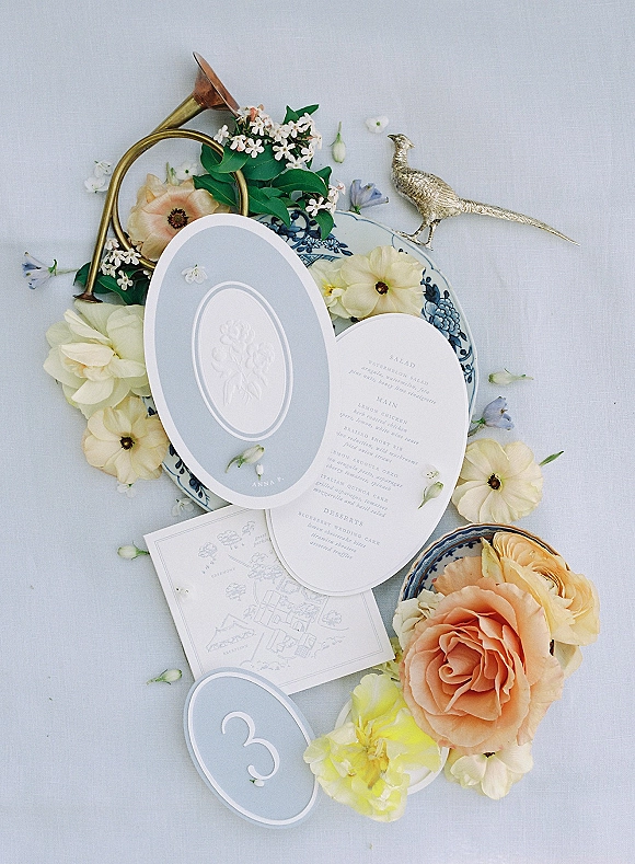 Invitation suite wedding invitation flat lay with calligraphy, wax seal, floral liner, porcelain plate and peach roses on light blue linen tablecloth
