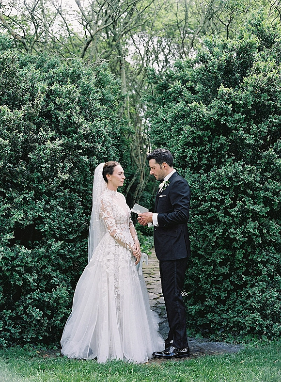 Wedding vows as groom reads from a vow book to bride in lace dress and veil, standing on a stone path by garden hedges