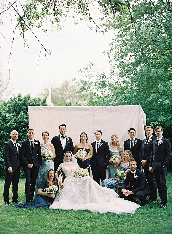 Wedding party portrait with bride seated in lace gown and long veil, bridesmaids in blue and groomsmen in black tuxedos on a garden lawn