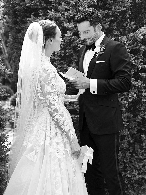 Wedding vows as the bride and groom vows from vow books, her lace long-sleeve dress and veil beside his tux, in garden greenery