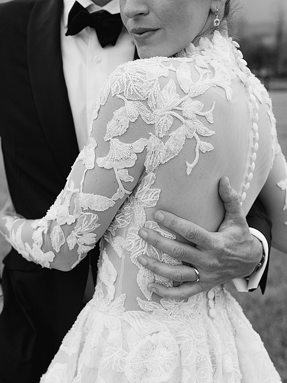 Wedding couple portrait of bride and groom embrace, close up on his hand at her waist showing lace sleeve detail in soft outdoor blur