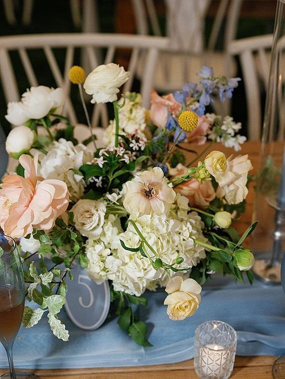Wedding centerpiece with hydrangea and roses, pastel wildflowers and greenery on a blue runner, with votive candle and table number on wood table