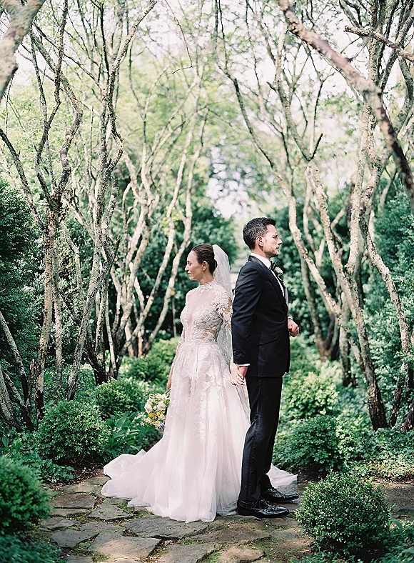 First look moment of bride and groom holding hands back to back on a tree-lined garden path, her veil and bouquet beside his black suit