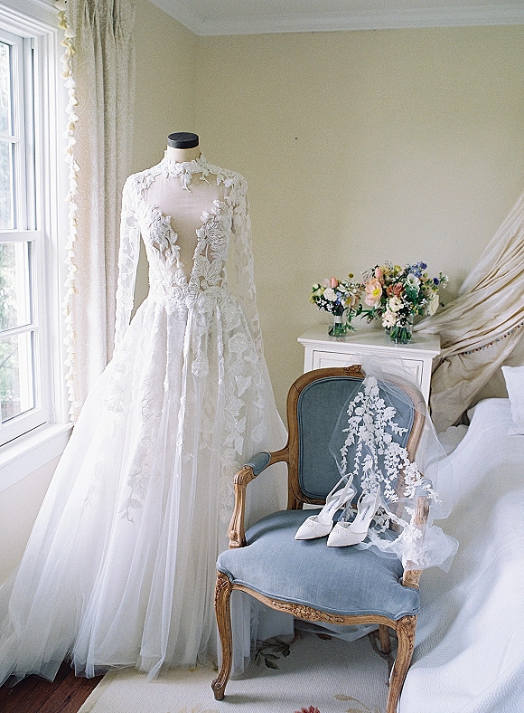 Wedding dress with long sleeve lace bodice and illusion neckline on an upholstered chair by a bedroom window with veil, bouquet, and heels