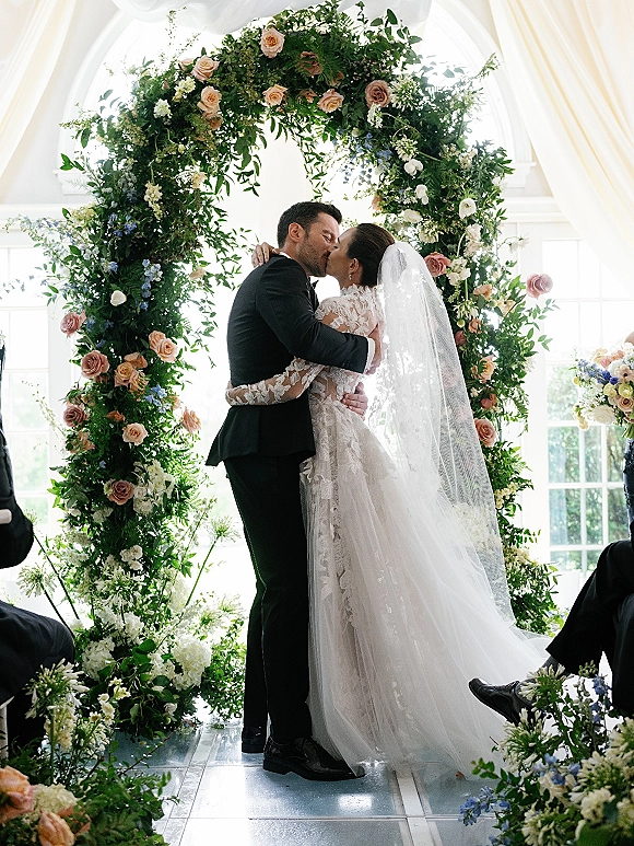 Wedding kiss as bride in lace dress and veil embraces groom in tux beneath a round floral arch with greenery, roses, and window light