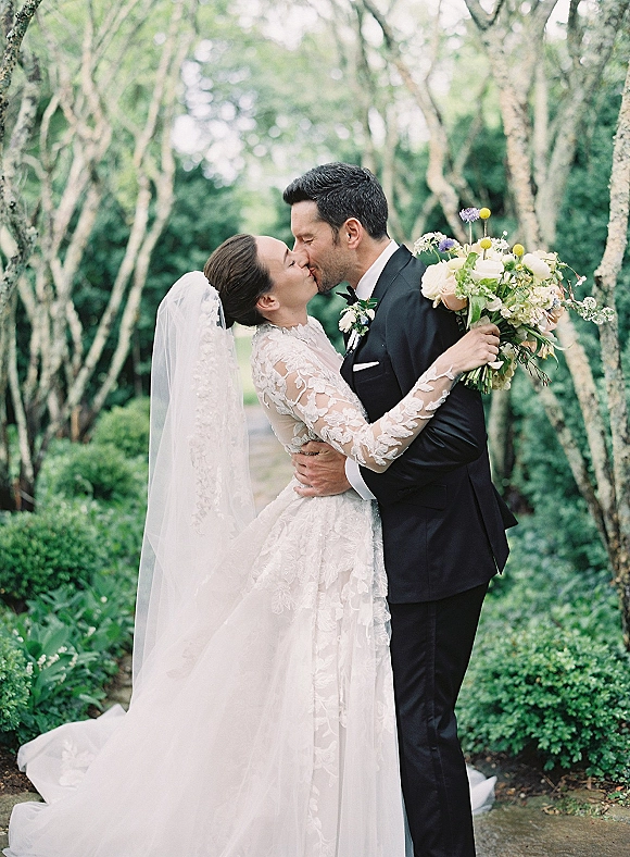 Wedding kiss portrait of bride and groom kissing on a tree-lined garden path, bride in lace sleeves and cathedral veil holding bouquet