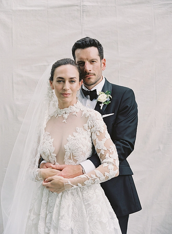 Couple portrait of groom hugging bride in a long sleeve lace wedding dress and cathedral veil against a neutral studio backdrop, black tuxedo bow tie
