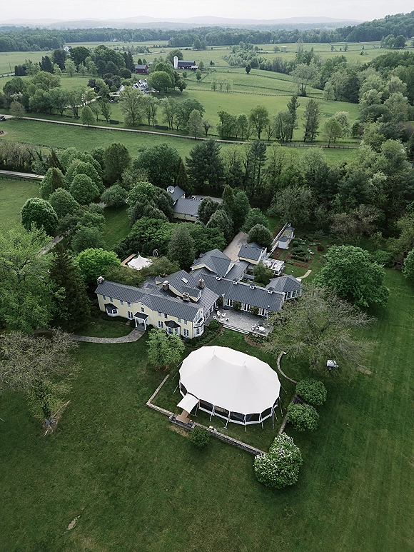 Outdoor wedding tent with wedding tent setup beside an estate house, white sailcloth canopy on green lawn amid pathways, shrubs, rolling hills
