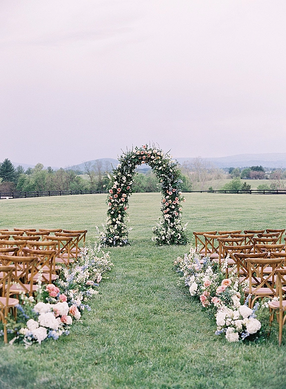 Ceremony setup with a floral ceremony arch and aisle flowers, featuring blush, white, and blue blooms on a grassy lawn with rolling hills