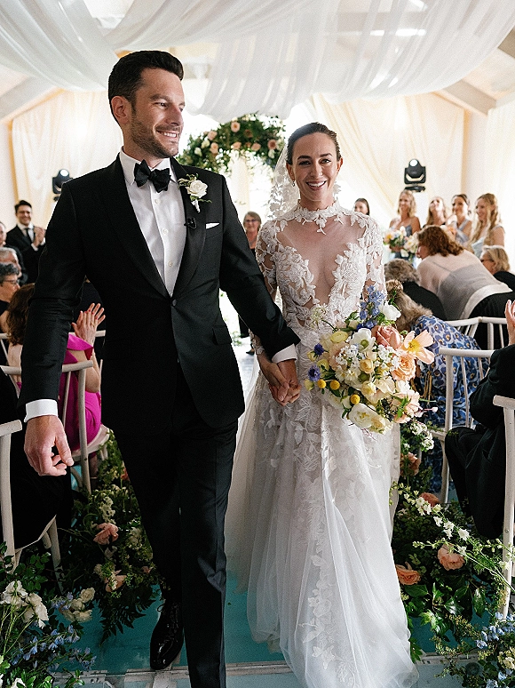 Wedding recessional as bride and groom walk hand in hand down an indoor aisle, lace gown and veil, guests cheering under floral arch