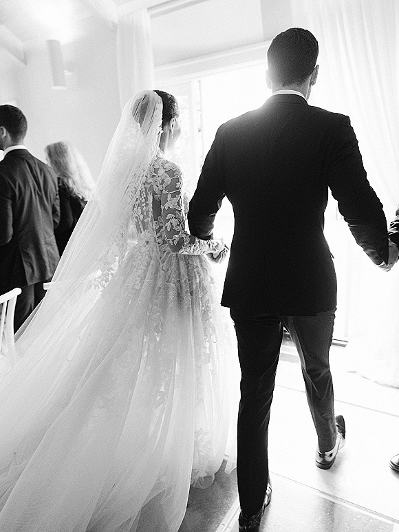 Wedding recessional of bride and groom walking away hand in hand, lace dress with long veil and train, guests lining an indoor aisle by a bright doorway