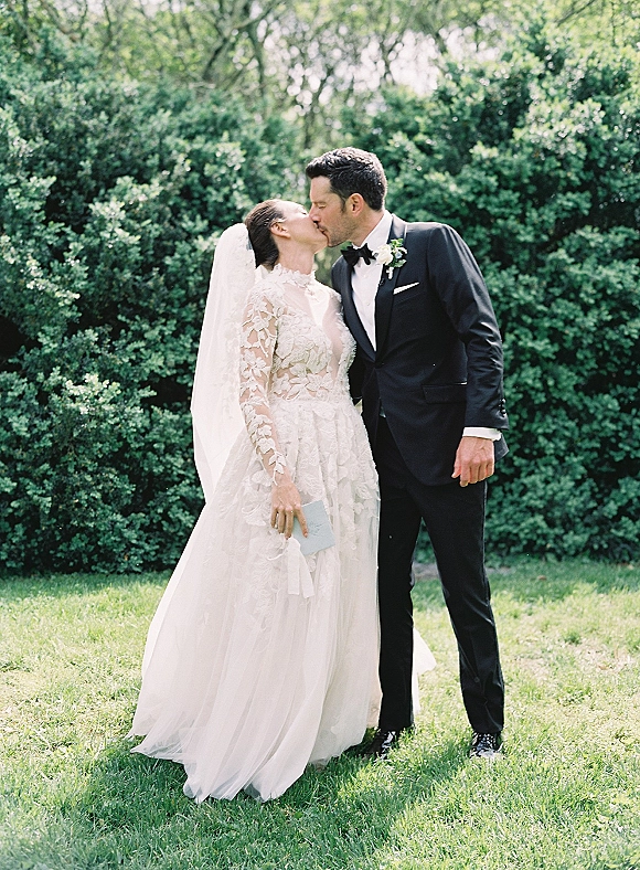 Wedding kiss portrait of bride and groom kissing outdoors, bride in lace long sleeves and veil beside groom in tuxedo against green hedges