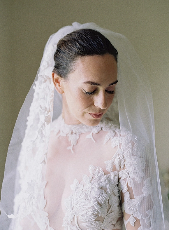 Bridal portrait of a bride looking down in a lace wedding dress with illusion neckline, floral appliqués, and veil against a neutral wall