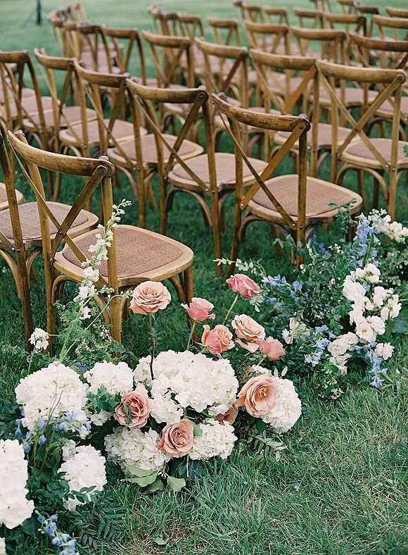 Ceremony aisle decor with outdoor ceremony aisle flowers of hydrangeas, blush roses, and greenery lining wood cross-back chairs on grass lawn