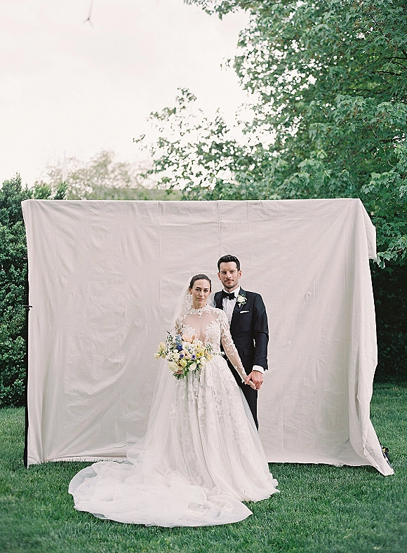 Couple portrait of bride and groom holding hands, her long veil and lace bodice beside his black tuxedo, on a lawn with fabric backdrop