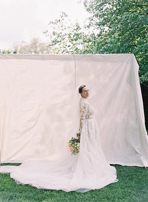 Bridal portrait of a bride in a long sleeve lace wedding dress holding a bouquet, cathedral veil and train flowing by a white fabric backdrop