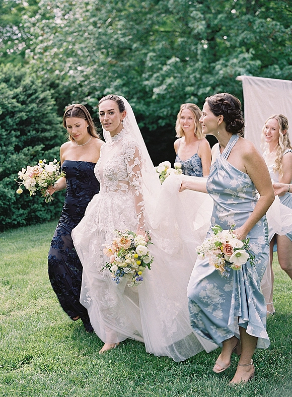 Bride with bridesmaids walking together, holding bouquets, as one adjusts her veil; lace-sleeved dress on a green lawn with trees behind