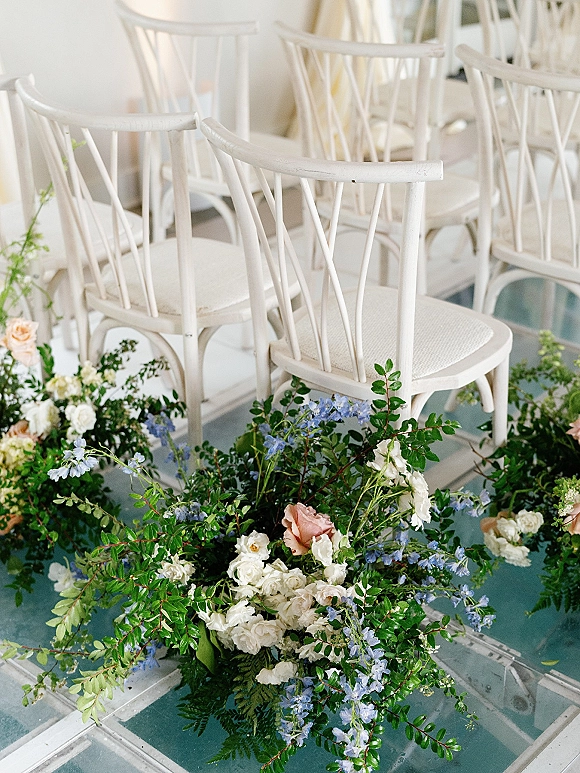Ceremony aisle flowers in a floor floral installation of white and peach roses with blue delphinium and ferns beside white wooden chairs indoors