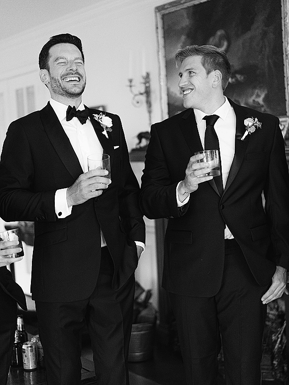 Groomsmen portrait of men in black tuxedos laughing with whiskey glasses, wearing boutonnières in an indoor room with framed painting