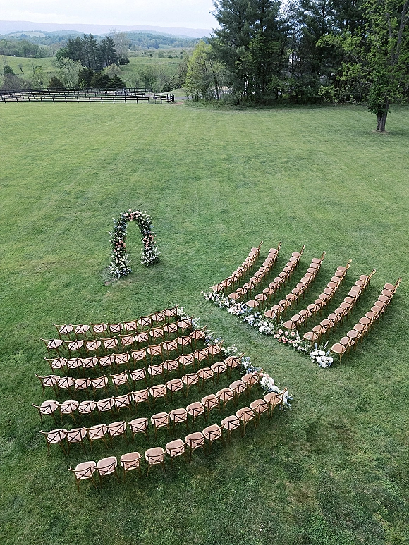 Outdoor ceremony setup with ceremony seating layout framing a curved aisle, floral arch, and rose greenery on a hilltop lawn by a wooden fence