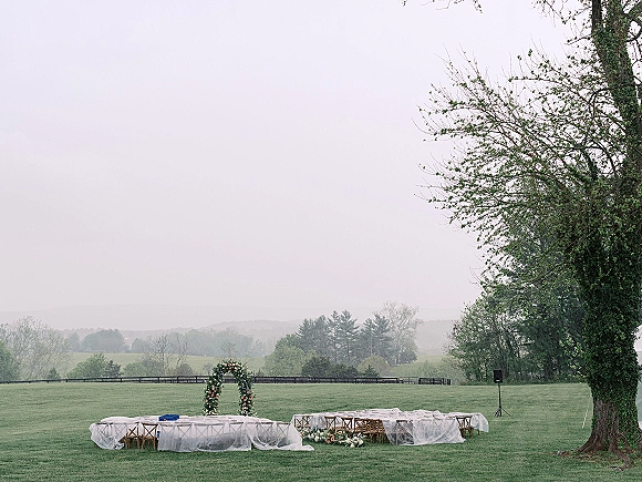 Outdoor reception setup with long banquet tables and crossback chairs under clear plastic covers on a grassy lawn with floral arch, trees, hills